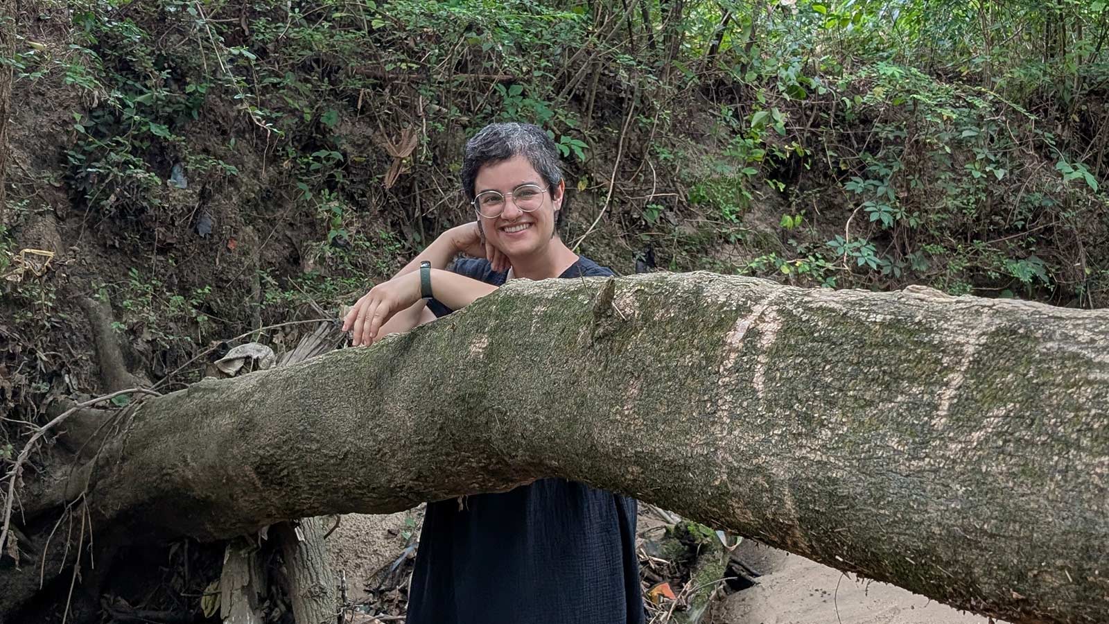 woman with short brown hair standing in creek and leaning on a fallen tree